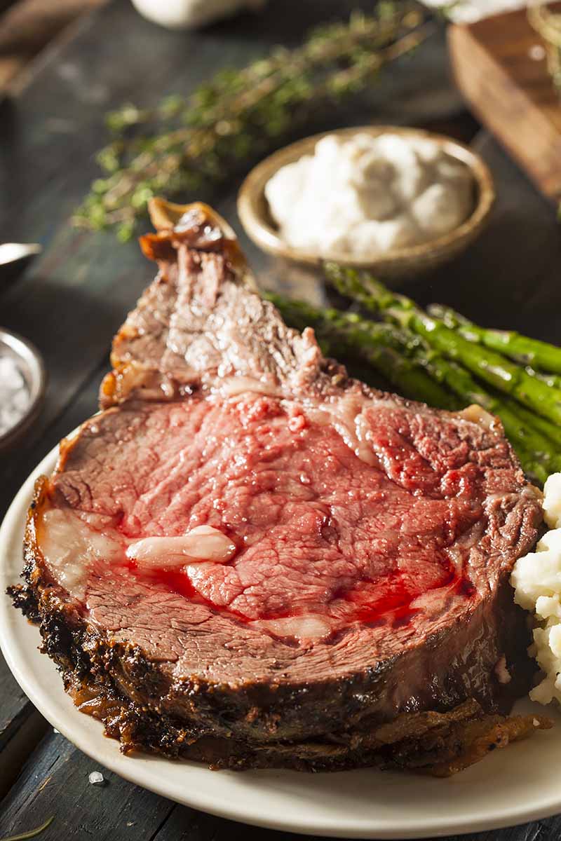 Vertical image of a slice of rare meat on a plate with mashed potatoes and asparagus next to sauces.