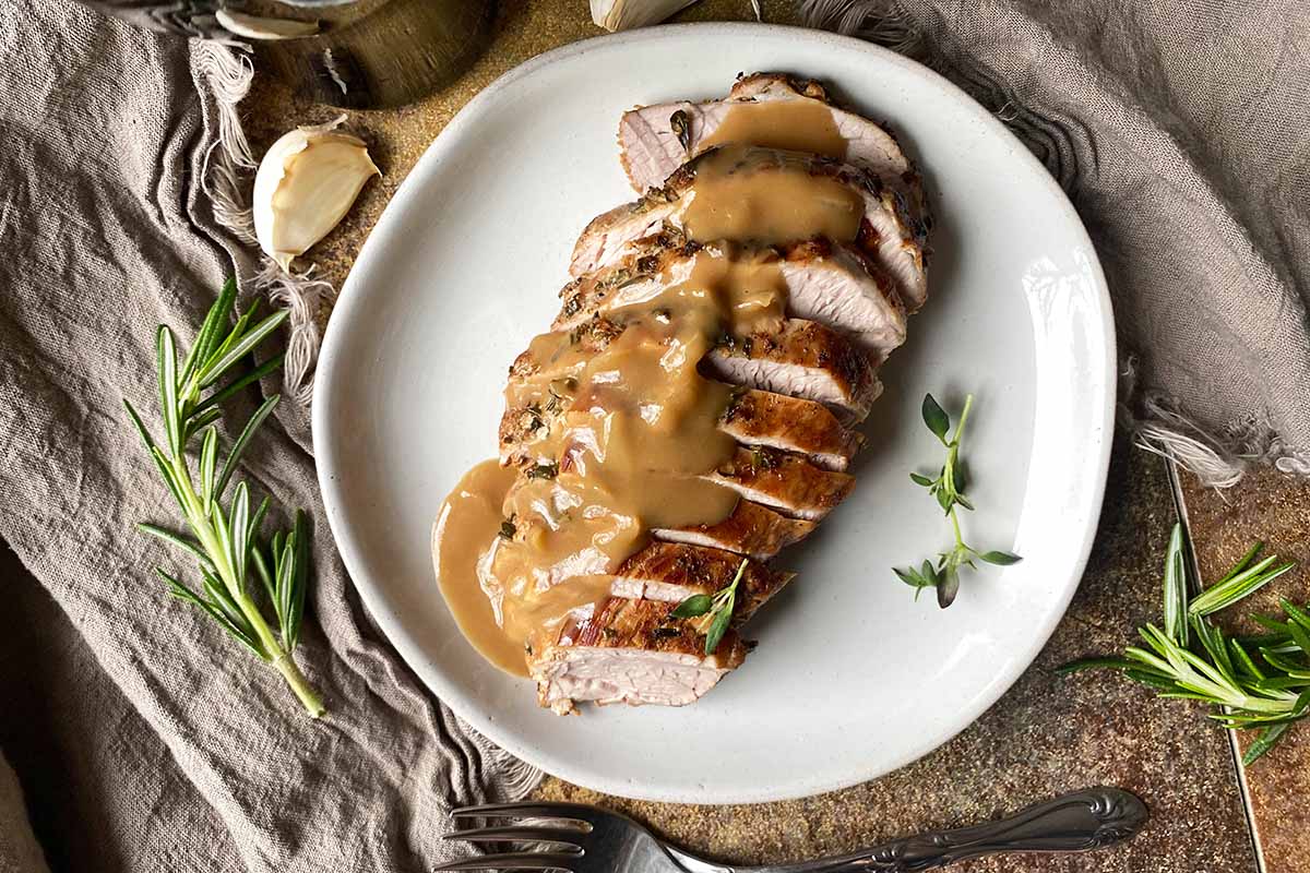 Horizontal top-down image of seared pieces of meat shingled on a white plate and drizzled with gravy next to tan towels.