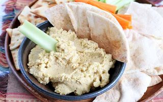 An appetizer platter of crudite, pita bread and chips, and a small navy blue bowl of beige garlic hummus, with a celery stick dipped into the spread.