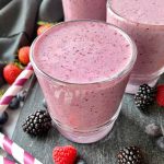Horizontal image of glasses filled with a bright purple beverage on a slate next to straws and fresh fruit.