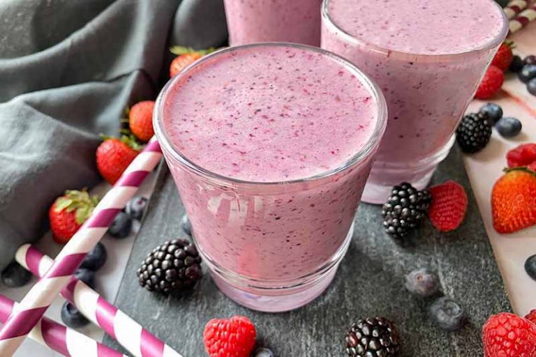 Horizontal image of glasses filled with a bright purple beverage on a slate next to straws and fresh fruit.