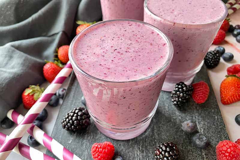 Horizontal image of glasses filled with a bright purple beverage on a slate next to straws and fresh fruit.