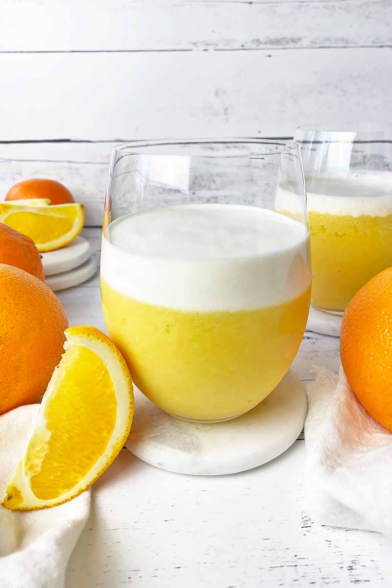 Vertical image of two glasses filled with a colorful shake topped with a white layer next to fresh fruit.
