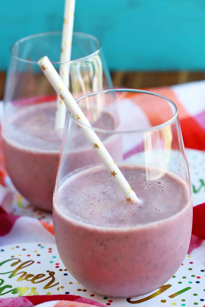 Two stemless wine glasses containing a pink smoothie with white paper straws printed with gold metallic stars, on paper decorative coasters on top of a red and white checkered cloth, on a brown wood surface with an aqua painted backdrop.