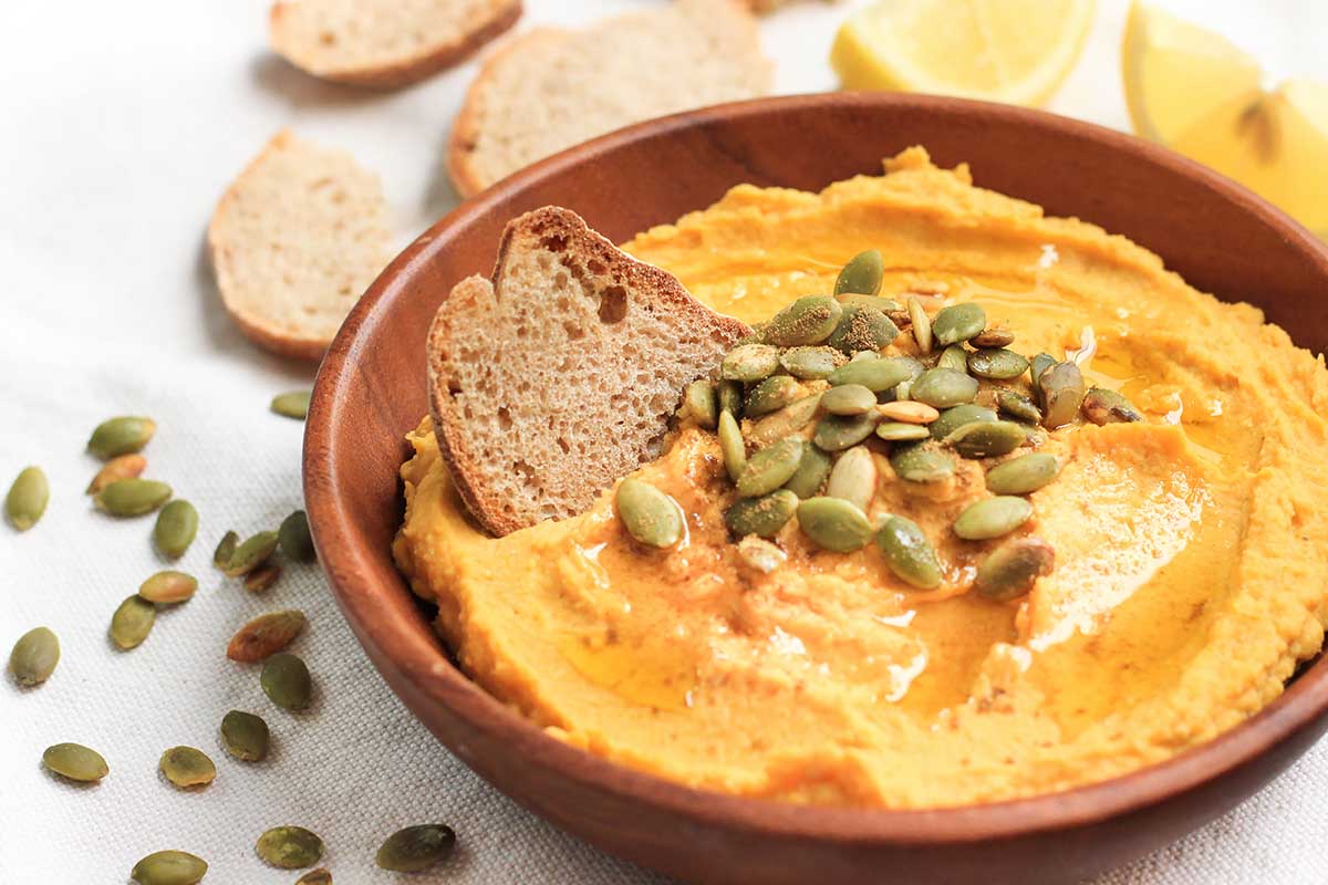 Horizontal image of a pumpkin dip in a wooden bowl next to seeds, slices of bread, and lemon wedges.