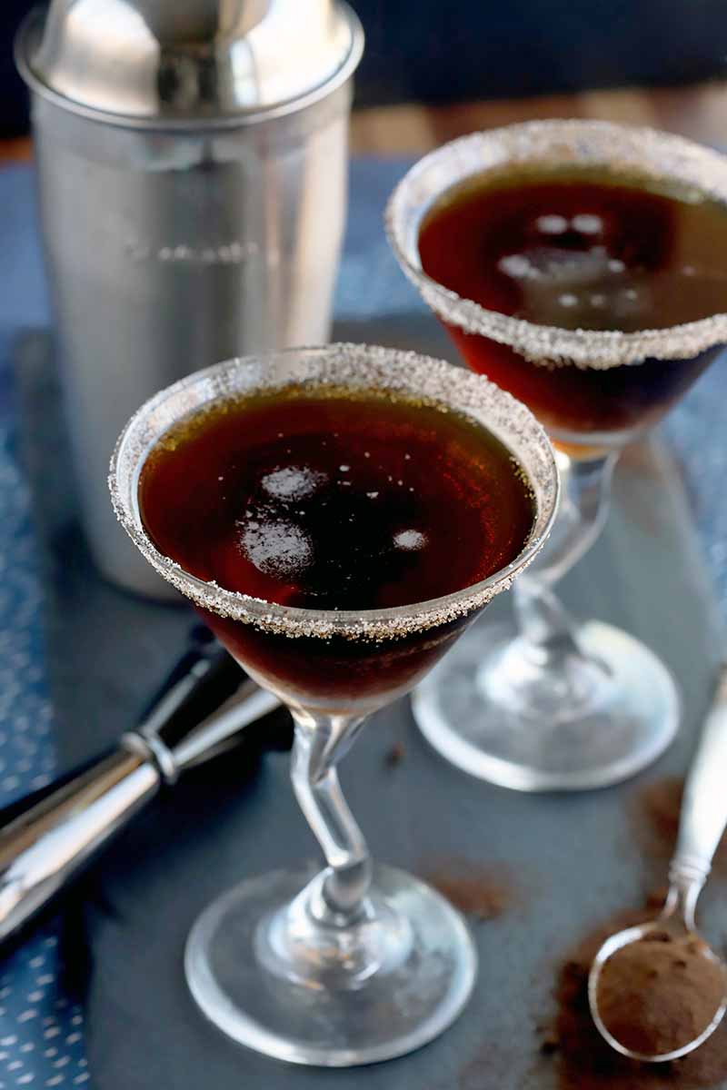 Vertical image of two coffee cocktails in martini glasses, with a stainless steel shaker and jigger, and a spoonful of coffee powder with more scattered around it on a slate serving board, on a blue and white cloth on a wood surface, with a charcoal gray background.
