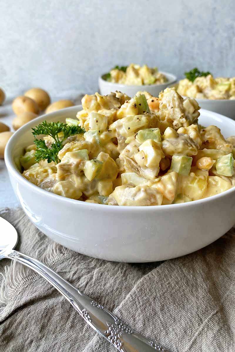 Vertical image of a large bowl with potato salad garnished with parsley on a tan towel with a metal serving spoon.