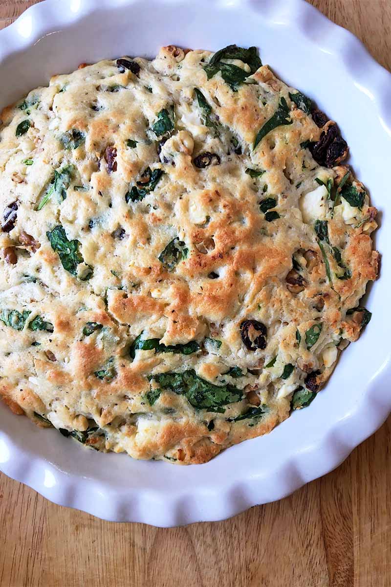 Vertical top-down image of a white pie plate with a baked good studded with spinach, nuts, and dried fruit on a wooden table.