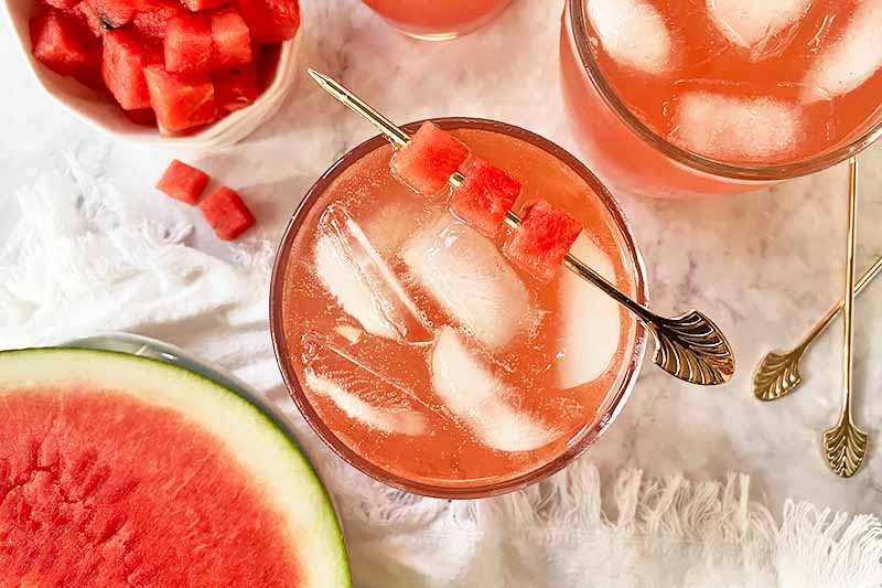 Horizontal top-down image of cups filled with a light pink liquid with ice cubes and a skewer of fruit cubes.