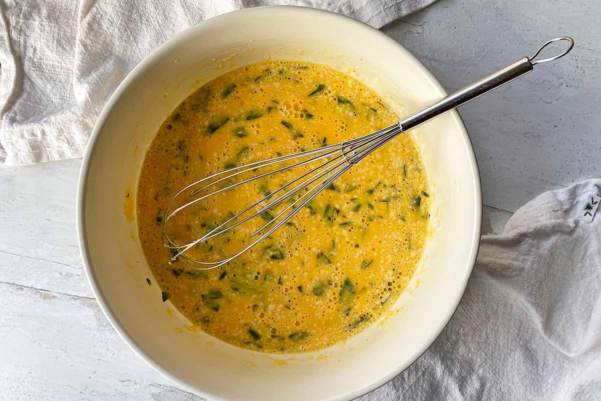 Horizontal image of whisking a liquid with chopped basil in a large white bowl.