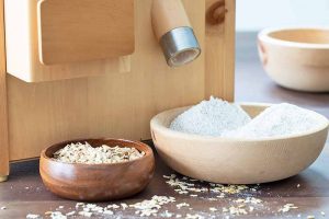 Horizontal image of whole and ground oats in bowls next to an appliance on a countertop.
