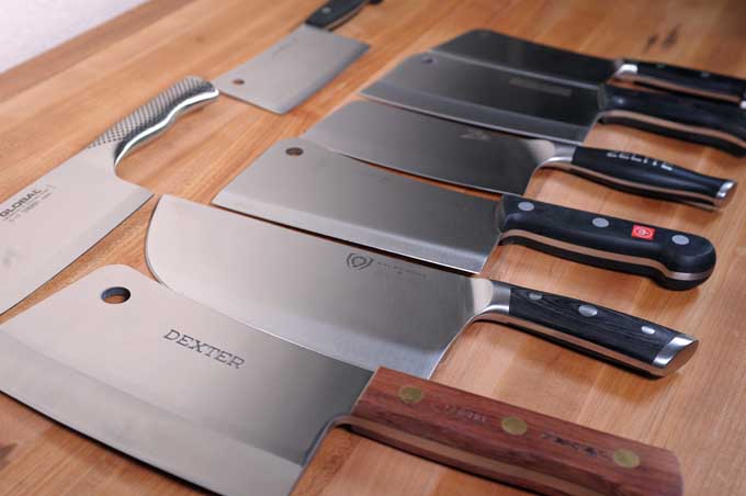 Oblique profile of eight different top rated meat cleavers on a wooden prep table.