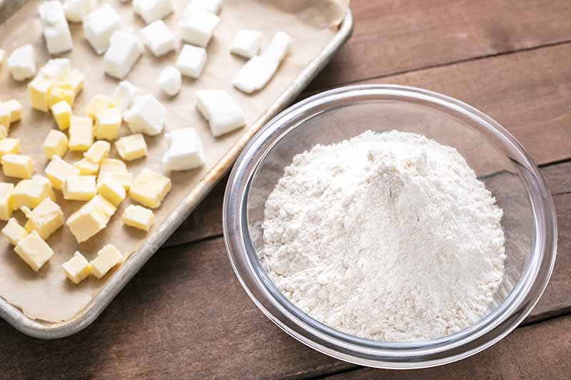 Horizontal image of a glass bowl with flour next to a baking sheet with cubed butter and shortening.