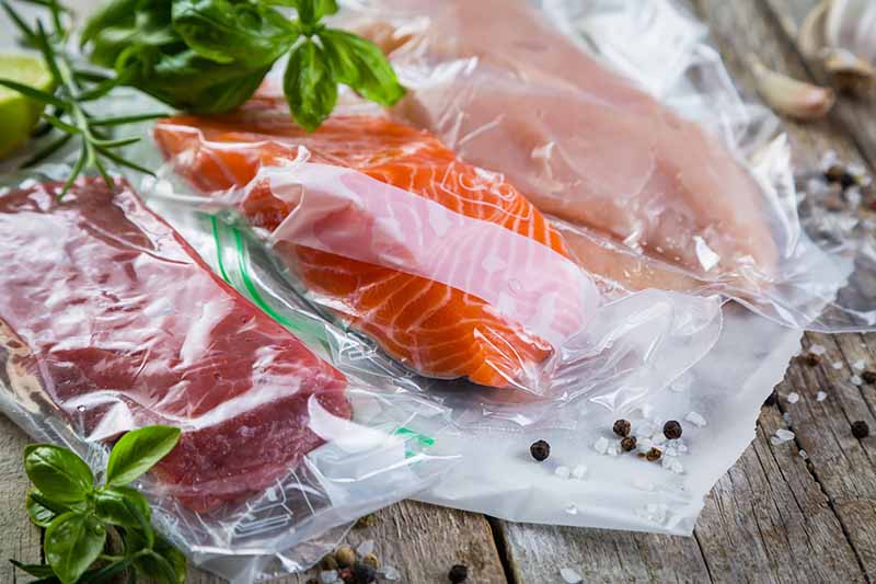 Horizontal image of cuts of raw meat in plastic next to herbs.