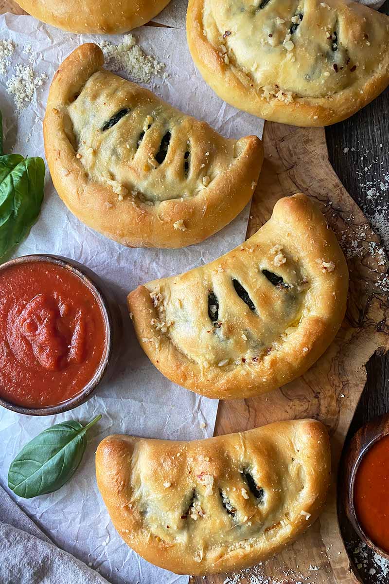 Vertical top-down image of small half-moon pastries with slits and doughy crusts on a wooden cutting board on top of parchment paper next to tomato sauce in bowls.