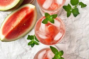 Horizontal top-down image of three glasses filled with a beverage with small balls of fruit, ice cubes, and fresh herb leaves next to white towels.