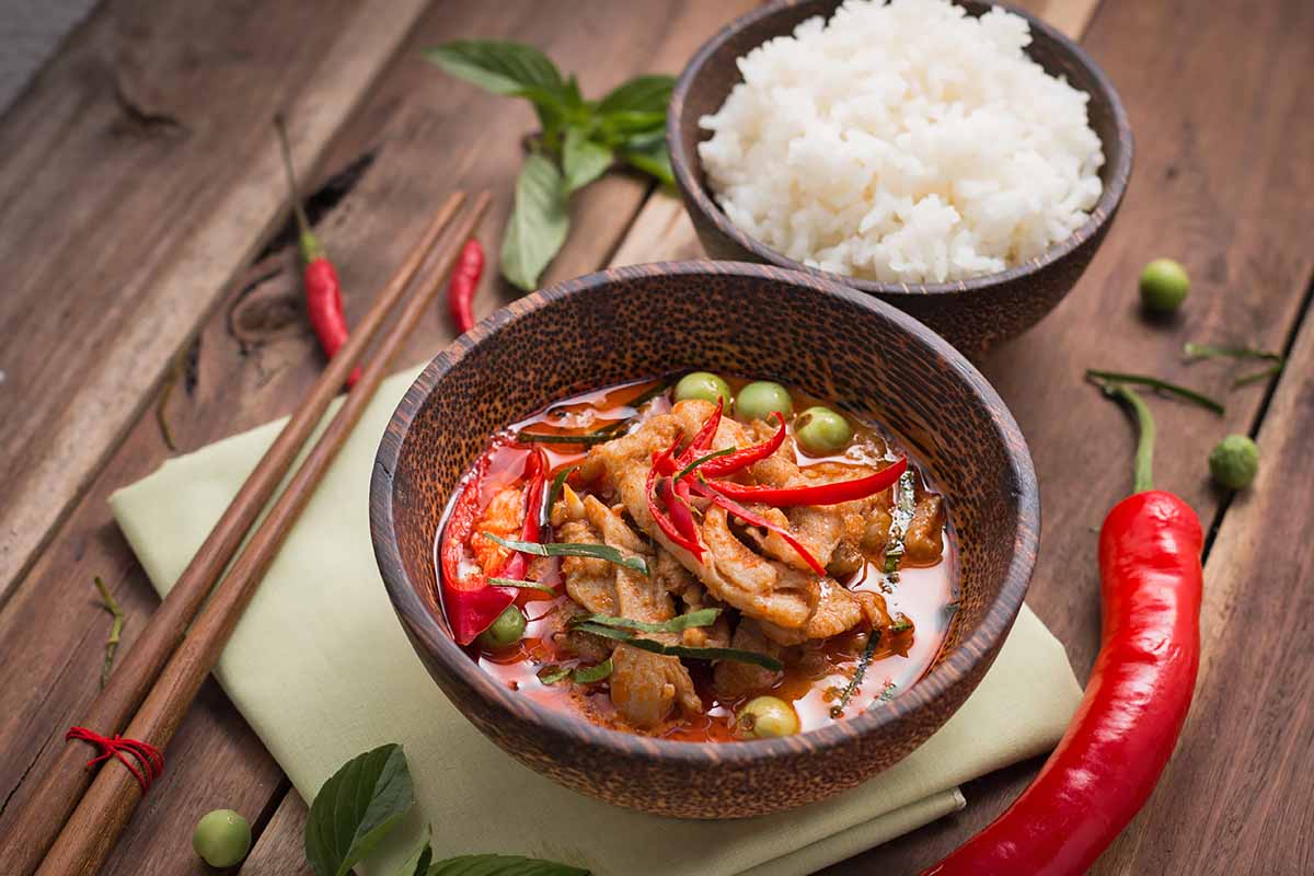 Horizontal image of a chicken curry in a bowl next to another bowl filled with rice, with chopsticks on the side.